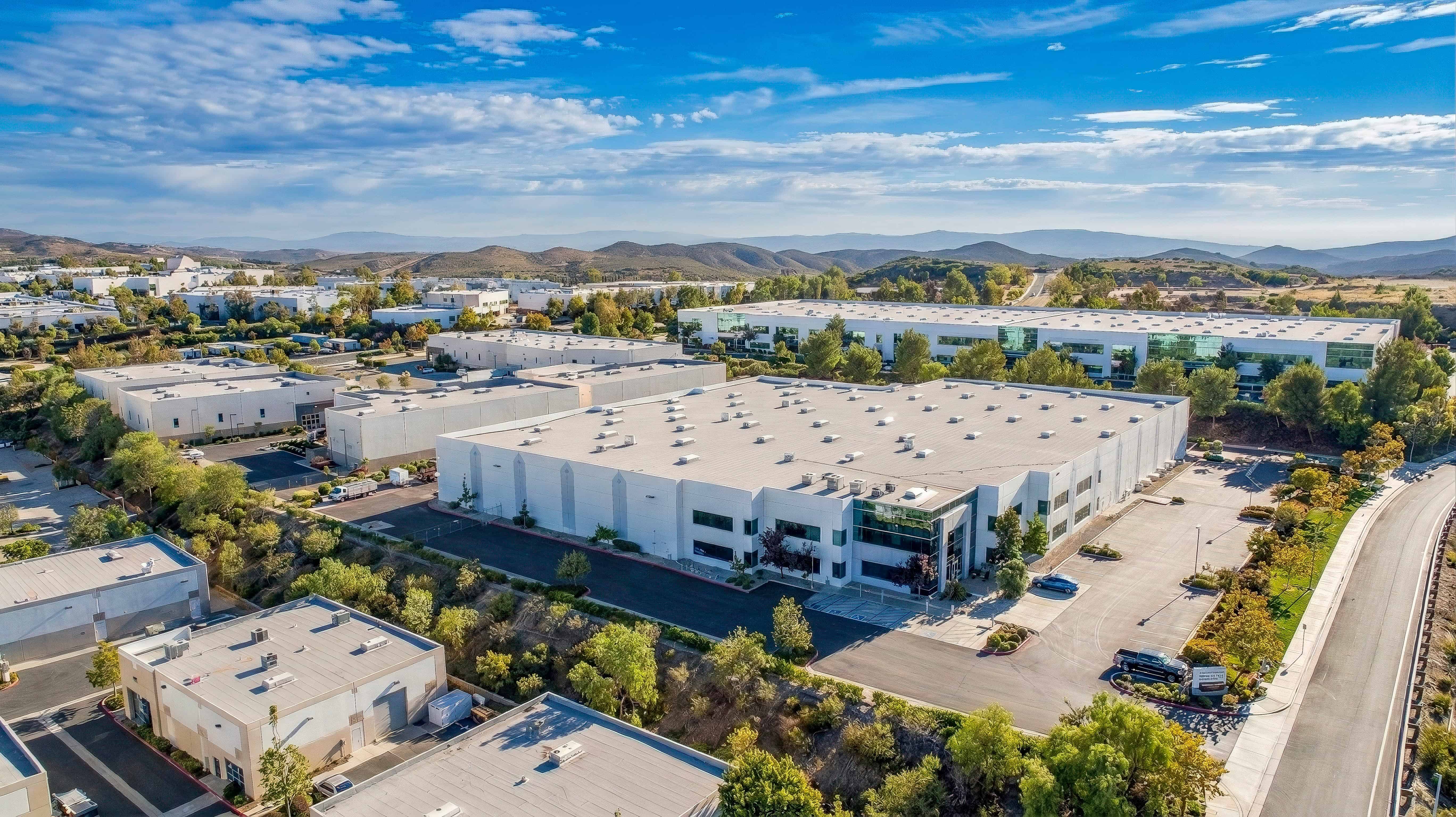 Large industrial warehouse with surrounding parking and greenery under a blue sky.