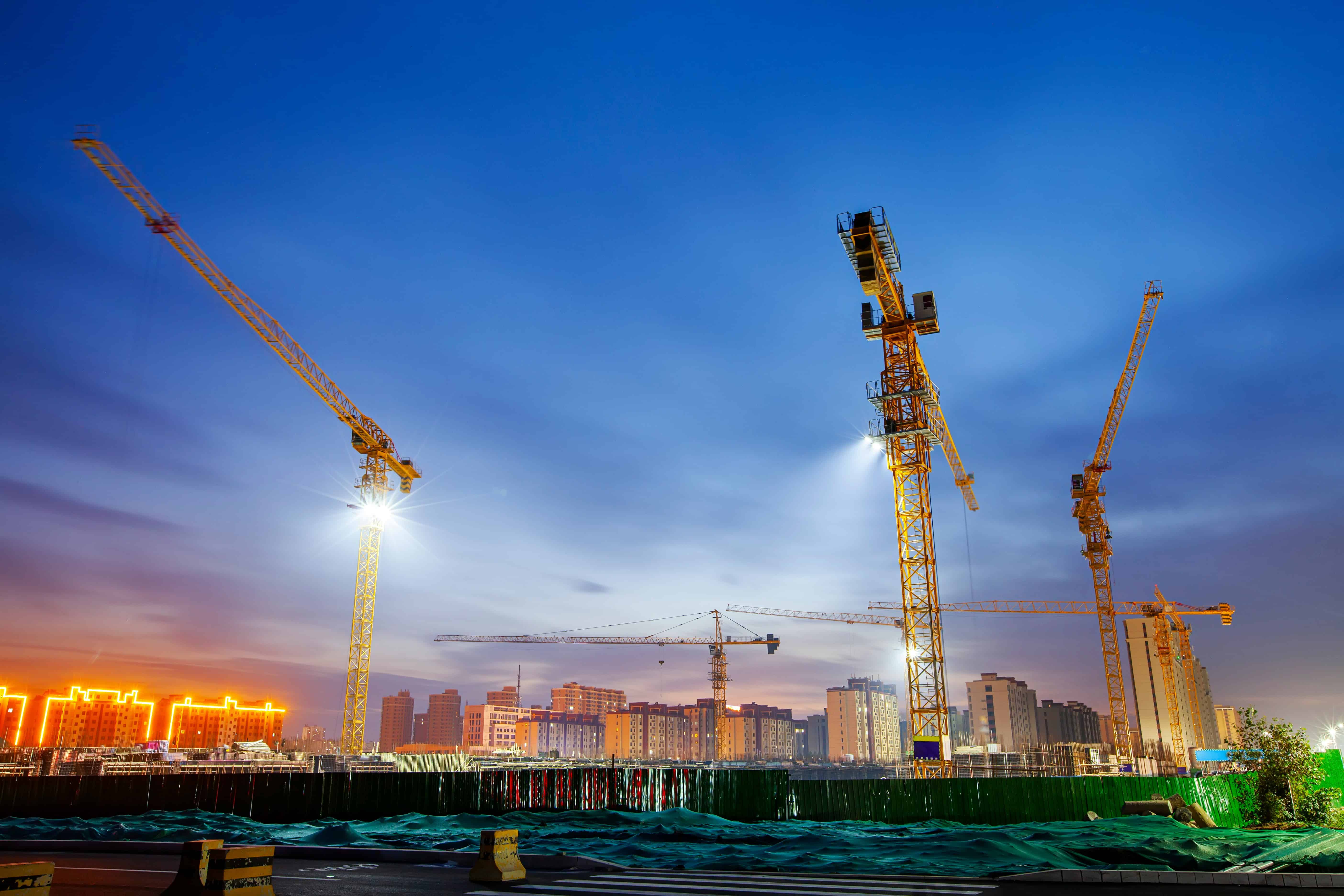 Construction cranes at work on a city skyline during sunset.