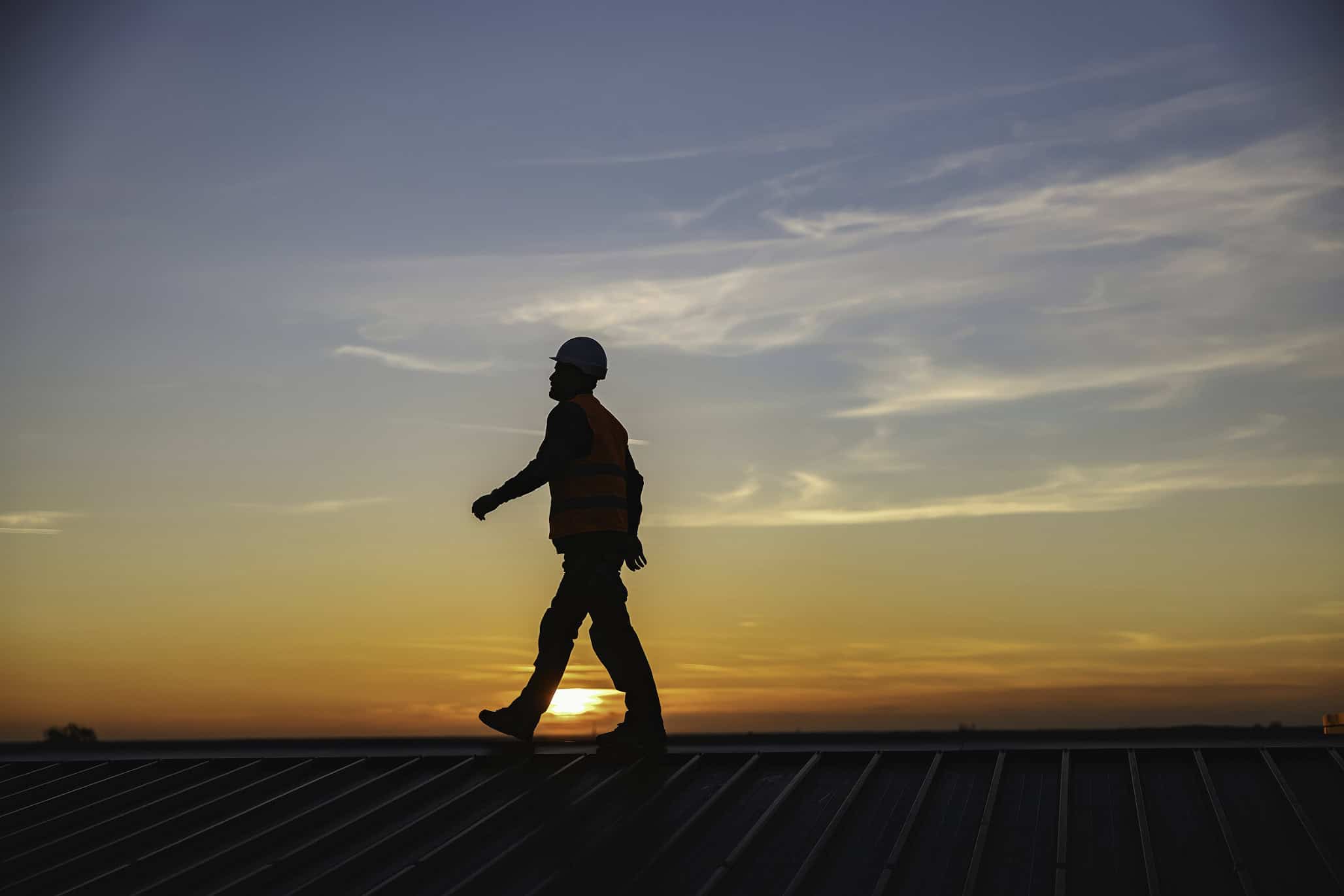 Construction worker walking on roof at sunset with safety gear.